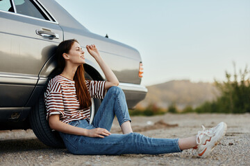 A young woman sits on the ground near her car on the side of the road and looks at the sunset