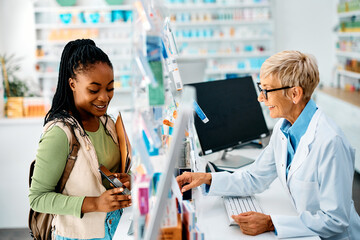 Young African American woman using smart phone while talking to pharmacist in drugstore.