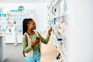 Fototapete Apotheke Junge schwarze Frau, die Handy benutzt, während sie in der Apotheke nach Medikamenten sucht.