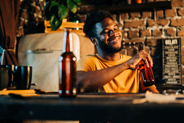 Obraz premium Shot of attractive african young happy smiling man drinking beer bottle in the bar close up portrait.