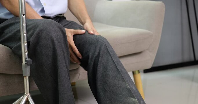 Senior grandfather with walking stick trying to exercise knee and leg on sofa at home, Elderly man with knee joint pain and suffering