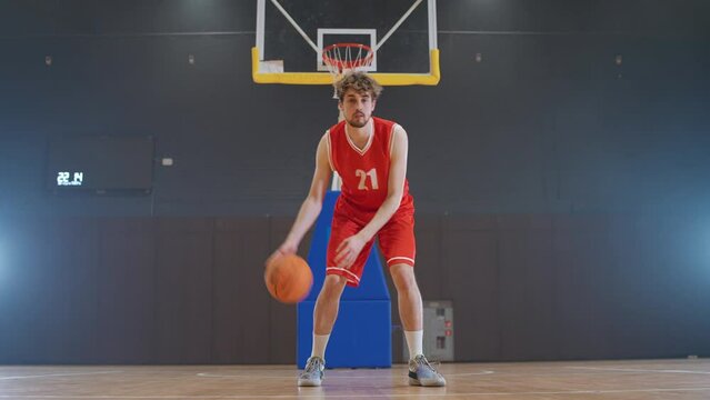 Basketball player hits the ball on the floor, warms up before the game, caucasian player looks at the camera, indoor playground.