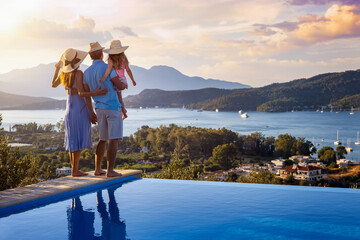 A family on summer holidays stands by the swimming pool and enjoys the beautiful sunset behind the...