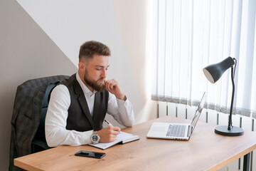 Business planning. Thoughtful bearded man using laptop and writing in notebook while sitting at table in office at work in business clothes, empty space