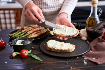 Making breakfast toasts - woman smearing cream cheese on a grilled bread over kitchen table with Asparagus wrapped with bacon and spices on a plate