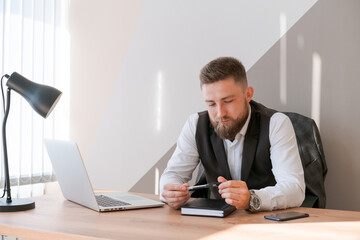 Handsome businessman is working with laptop in office, in business clothes at his desk. A bearded successful guy does his job perfectly well. Reflects and analyzes his actions