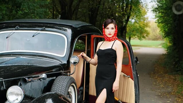 Young elegant retro sexy woman in black evening dress, red stylish vintage scarf on head stands near old style retro car. road rain green grass summer nature park tree. Girl driver fashion model lady
