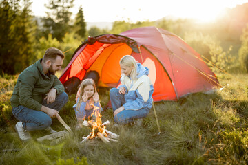 Young adult couple sit with a little girl by the campfire at campsite, traveling with tent on nature. Family spend summer time hiking in the mountains © rh2010