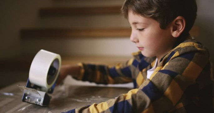 Child, moving and new house while packing a cardboard box for relocation to his new foster care home. Adoption with a young child using clear tape for a package while helping pack for a fresh start