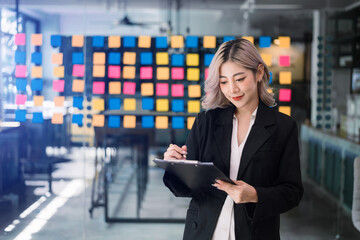 Confident Asian woman with a smile standing holding notepad and tablet at the modern office..