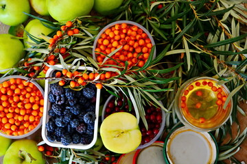 Berry and fruit background with autumn stocks still-life. Sea buckthorn, bird cherry, blackberries in jars and cup of berry tea between twigs and ripe green apples. Top view, flat lay 