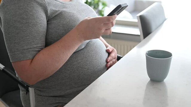 Pregnant woman in casual clothing using smart phone at kitchen table in the morning. Coffee cup standing on the table. Footage made in Sweden.
