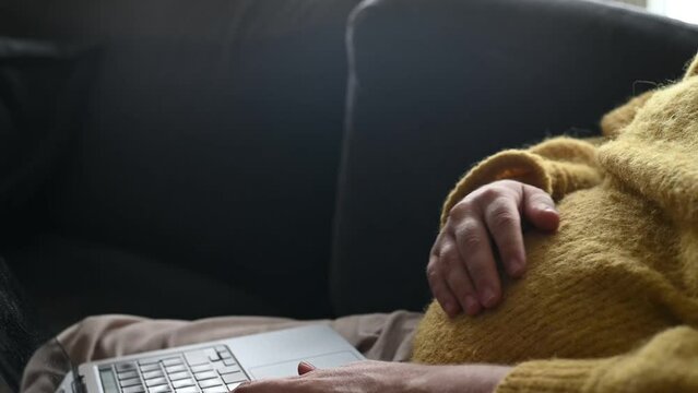 Pregnant woman working from home using laptop. Expecting woman sitting in the sofa indoors in a warm yellow sweater, working remote and typing on laptop keyboard.