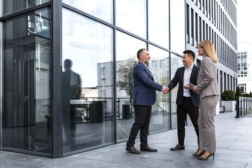 Meeting of three successful business people, diverse dream team man and woman outside office building, greeting and shaking hands, experienced professionals specialists in business suits talking