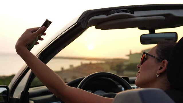 Attractive woman makes selfie and takes photos of the sunset from her convertible car parked in front of the beach, enjoying the sunset alone and relaxing with the view