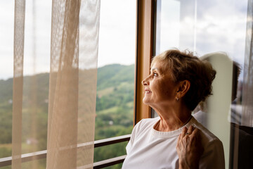 Beautiful happy gray-haired mature woman in a white t-shirt standing by the window enjoying the morning and nature