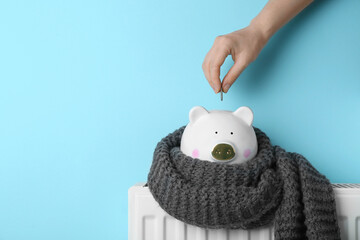 Woman putting coin into piggy bank on heating radiator against light blue background, closeup. Space for text