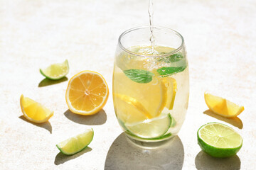 Pouring water into glass with lemon slices and mint at light grey table