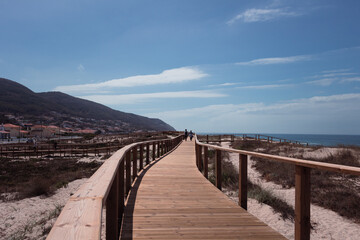A great spot for a pleasant walk by the sea on a warm summer afternoon. Long wooden pier boardwalk and handrail in Quiaios beach with distant houses, mountain and the blue sky above
