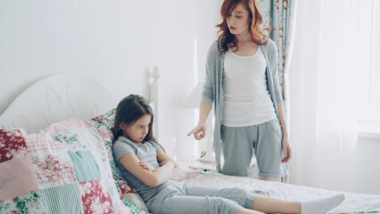Angry mother argue scolding her upset little daughter lying on bed in cozy bright bedroom at home