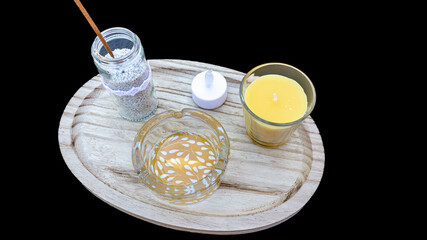 Simple decorative centerpiece against a black studio background, glass ashtray with white leaves, yellow candle in a holder, incense in a jar with white pebbles and a small lamp on a wooden plate