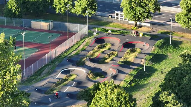 Young people on scooters and mountain bikes ride a hilly skate park next to the tennis courts in Hillsborough Park in Sheffield on a sunny summer day. Drone dolley tilt shot