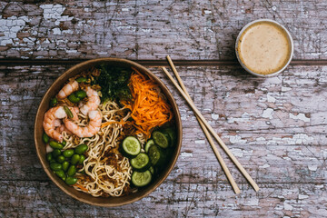 Noodle Poke Bowl with Shrimps, Edamame, Wakame Seaweed, Shredded Carrots, Cucumber and Fried Onion bites near to a cup of Hot Cajun Sauce and a pair of Chopsticks over an Old Wooden Texture