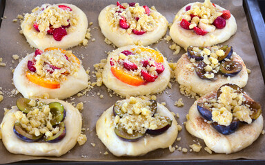 Risen, unbaked sweet rolls (yeast dough) with various seasonal fruits (strawberries, nectarines, plums, bananas) sprinkled with crumble. The rolls lie on a baking tray lined with paper. Top view.