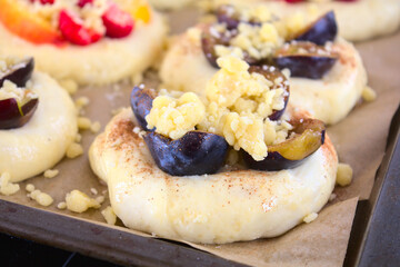 Close-up of a risen, unbaked sweet roll (yeast dough) with plums, cymon and crumble. In the background, rolls with seasonal fruit on a baking tray lined with paper. Shallow depth of field.