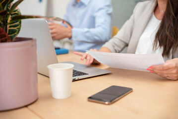 Close-up of unrecognizable businesswoman sitting at the table in front of laptop and holding a document in her hand