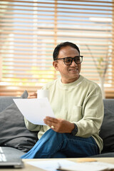 Happy senior man holding paper, make loan insurance payment on laptop while sitting in cozy living room. Elderly technology concept