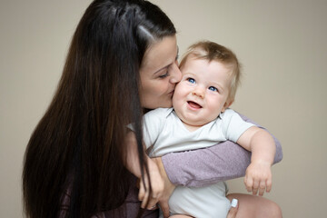 pretty young mother with dark hair, purple top is holding her 7 months old baby and standing in front of brown background and is happy and full of love