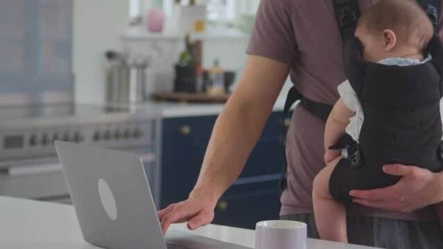 Close Up Of Transgender Father Working From Home On Laptop Looking After Sleeping Baby Son In Sling