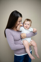 pretty young mother with dark hair, purple top is holding her 7 months old baby and standing in front of brown background and is happy and full of love