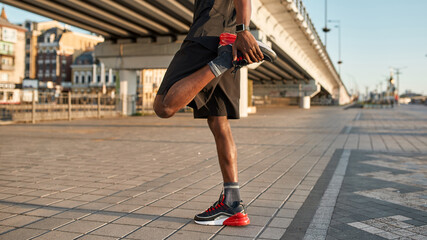 Cropped of black sportsman warming up leg in city - obrazy, fototapety, plakaty