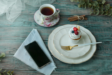 Vanilla cupcake with fruits, hot cup of tea and mobile phone on wooden table background, top view, copy space. Seasonal, morning Sunday breakfast and rest.