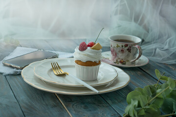 Vanilla cupcake with fruits, a hot cup of tea and a mobile phone near the window, on the background of a wooden table, top view, copy space. Seasonal, morning Sunday breakfast and rest.

