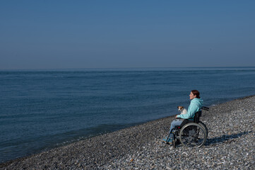Naklejka premium A serene Caucasian woman in a wheelchair is resting on the seashore with a jack russell terrier dog.