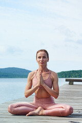 Fototapeta na wymiar mature woman in a pink tracksuit sits in a lotus position, practices yoga near the sea. Healthy lifestyle concept