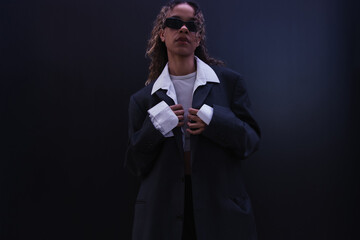 Obraz premium stylish african american woman in sunglasses adjusting oversize suit near dark blue wall.