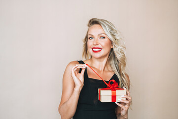 Portrait of gorgeous happy blonde young woman with bright makeup in evening dress with gift box in...