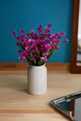 Close-up shot of a bouquet of realistic artificial orchids in a white vase. The vase of purple flowers stands on a wooden floor with a mirror on a blue background. Front view.