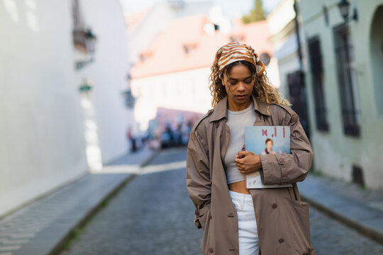curly african american woman in headscarf and trench coat walking with magazine on street in prague.