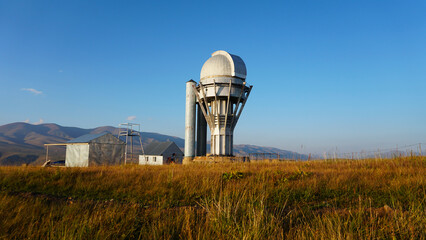 Large buildings of the observatory in the form of a dome. The telescopes are closed. Beautiful tall yellow-green grass develops in the wind. Blue sky and green hills in the distance. The Assy Plateau © SergeyPanikhin
