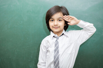 Portrait of happy schoolboy salute due to independence day or republic day while standing against chalkboard, He is wearing school uniform.