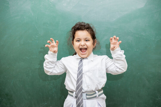 Little indian school girl kid with crazy expression standing against blank chalkboard or blackboard. Mad shouting student child doing fun in classroom.