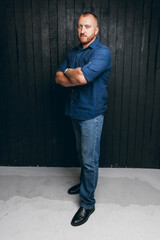 Portrait of a bearded man with a serious look, standing on a black wooden background. a business man in a blue shirt and jeans