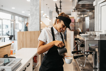 Beautiful and happy young female worker working in a bakery or fast food restaurant and using coffee machine. Positive people in small business concept. - obrazy, fototapety, plakaty