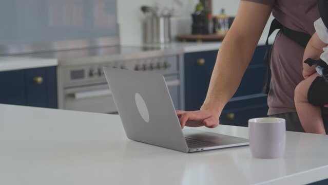 Close Up Of Transgender Father Working From Home On Laptop Looking After Baby Son In Sling
