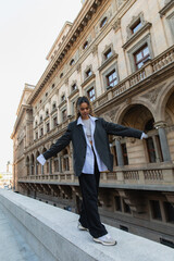 full length of curly african american woman in oversize suit walking with outstretched hands near building of national theater in prague.